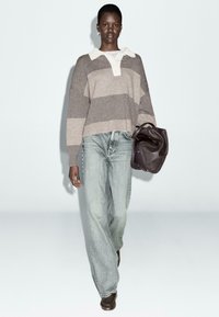 Pull rayé en taupe et crème, associé à un jean large bleu clair. Le mannequin porte un sac en cuir marron et des chaussures marron.
