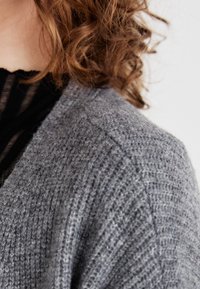 Lys grå strikket cardigan med en teksturert finish, med bred krage og senket skuldre, som delvis dekker en svart topp under.