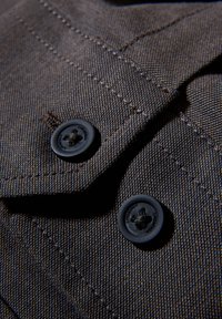 Brunt tyg med en texturerad väv, med två svarta knappar och synliga sömmar. Knapphål finns intill knapparna.