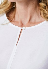 Top bianco a maniche corte con scollo rotondo, chiusura a bottone trasparente sul collo e una texture sottile, con un sottile bordo bianco lungo i bordi.