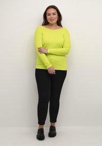 Dlouhý rukáv, žebrovaný top v jasné neonově žluté barvě, spárovaný s černými skinny džínami a černými polobotkami. Jednoduchý design s širokým výstřihem.