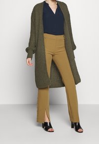 Una donna che indossa un cardigan lungo in maglia verde oliva, una blusa blu navy, pantaloni color cammello con fessure frontali e scarpe con tacco aperte nere.
