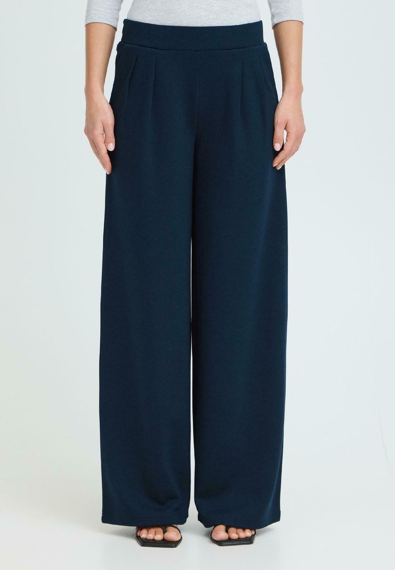Femme portant un pantalon large bleu marine et des sandales noires ouvertes, debout devant un fond clair uni.