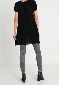 Tunique noire à manches courtes bouffantes, avec un tissu texturé et une silhouette fluide, portée avec un jean gris slim et des bottines.