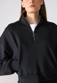 Black zip-up sweatshirt in a soft, smooth fabric. Features a high collar and drop shoulders, with a small metal zipper and an accent seam.