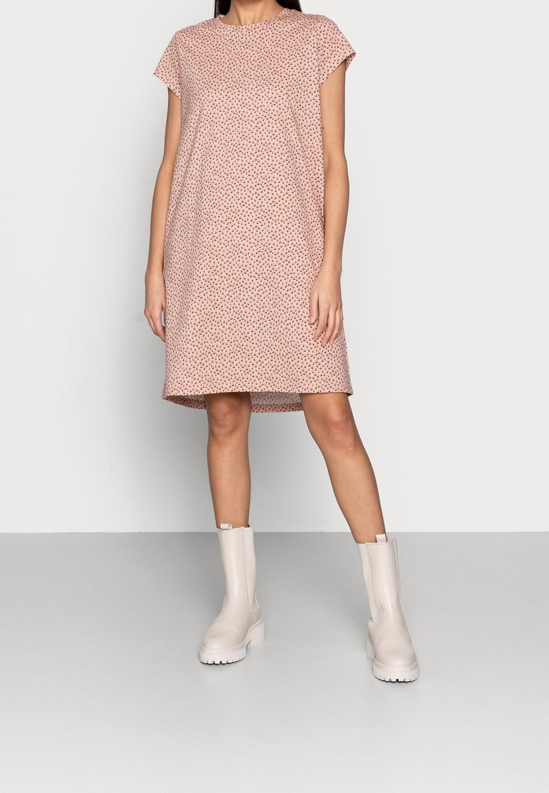 Femme portant une robe rose pâle à manches courtes, longueur genou, avec de petits motifs noirs, et des bottes blanches épaisses mi-mollet, debout sur un sol gris.