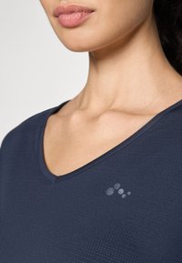 Marinblå atletisk topp med V-krage och en subtil texturerad design samt en diskret logotypdetalj på bröstet. Mjuk tygkvalitet och figurnära passform.