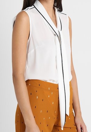Blouse blanche sans manches avec une longue cravate bordée de noir, associée à un pantalon orange taille haute avec un petit motif de tirets noirs et blancs.