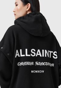 Must have suuruses musta kapuutsiga särk, kus seljaosas on suur valge tekst "ALLSAINTS". Lisaks on varrukatel erinevates fontides täiendav tekst ja nööbid.