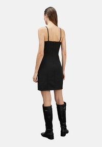Vestido negro ajustado con tirantes finos, que presenta una cremallera en la espalda. Acompañado de botas de cuero negras hasta la rodilla con herrajes decorativos.