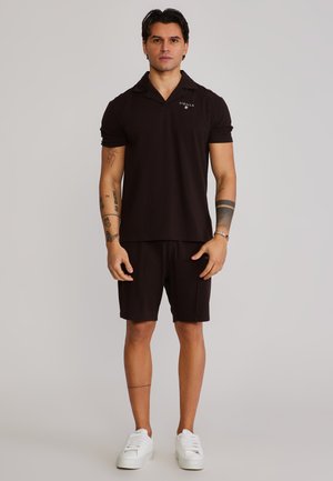 Mand står iført sort poloshirt og shorts med hvide sneakers, synlige tatoveringer på armen, mod en ensartet lys baggrund.