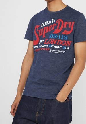 Mand iført en mørkeblå Superdry T-shirt med rød og hvid tekst, kombineret med mørkeblå jeans, hånd i lommen.
