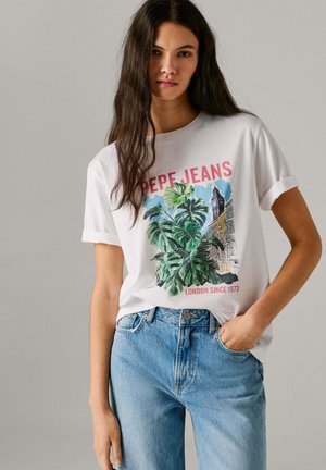 Jeune femme aux longs cheveux foncés portant un t-shirt blanc à motifs et un jean bleu, main dans la poche, debout devant un fond uni.