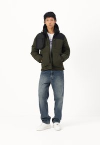 Pufjakke i olivengrøn med et sort skulderpanel, der har flere lommer. Båret med løse blå jeans og hvide sneakers.