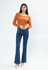 Oranges langärmliges Oberteil mit rundem Ausschnitt, kombiniert mit hochtaillierten blauen Schlagjeans, die goldene Knopfdetails an den Seiten haben.