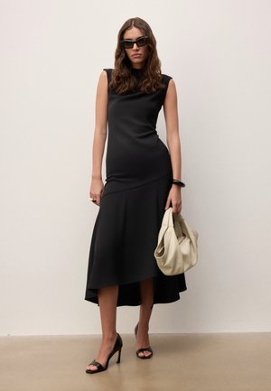 Femme en robe noire sans manches, sandales à talons noires, lunettes de soleil, tenant un sac à main beige sur un fond clair uni.