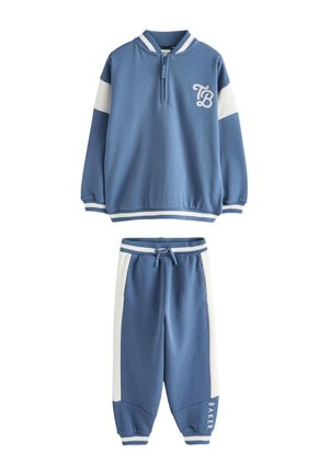 Niño pequeño con chándal azul y blanco y zapatillas blancas, de pie sobre un suelo gris, mirando hacia un lado con las manos juntas.