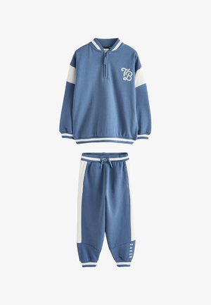Ung dreng iført blå og hvildt træningstøj og hvide sneakers, der står på en grå gulv og kigger til siden med hænderne foldet.