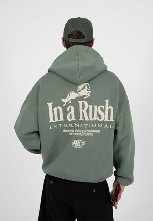 Person trägt grünen Hoodie und passende Mütze, auf dem Rücken ein weißes Pferde-Graphic und der Text "In a Rush International", hält eine Brieftasche.