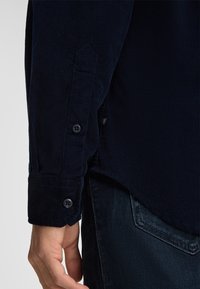Marineblaue Cordhemd mit geknöpften Manschetten und strukturiertem Stoff, mit einem dezent abgerundeten Saum und einem kleinen Logo-Tag an der Seite.
