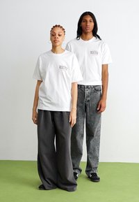 White cotton t-shirts with logo accents, worn by two models. One in gray wide-leg pants, the other in distressed denim, black sneakers.