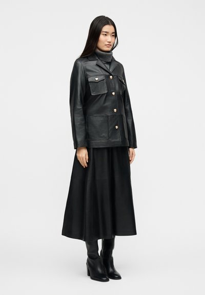 Γυναίκα που φοράει μαύρο δερμάτινο μπουφάν με χρυσά κουμπιά, μαύρη μίντι φούστα, μαύρες μπότες με τακούνι και γκρι πουλόβερ με ψηλό λαιμό, σε λευκό φόντο.