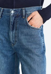 Mid-rise blue denim jeans featuring a straight leg, five-pocket design, contrasting orange stitching, and metal button closure.