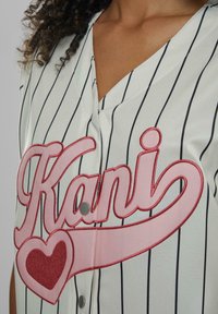 Hvit baseballtrøye med svarte striper. Har rød "Kani" tekst og hjerte-design, med knappestolpe og V-hals. Glatt tekstur.