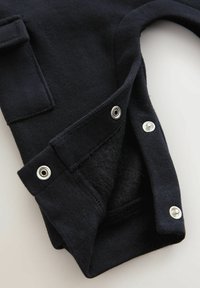 Schwarzes Kleidungsstück mit weicher Textur, ausgestattet mit Druckknöpfen, einer Taschenzier und einem runden Ausschnitt. Der Stoff wirkt warm und elastisch.