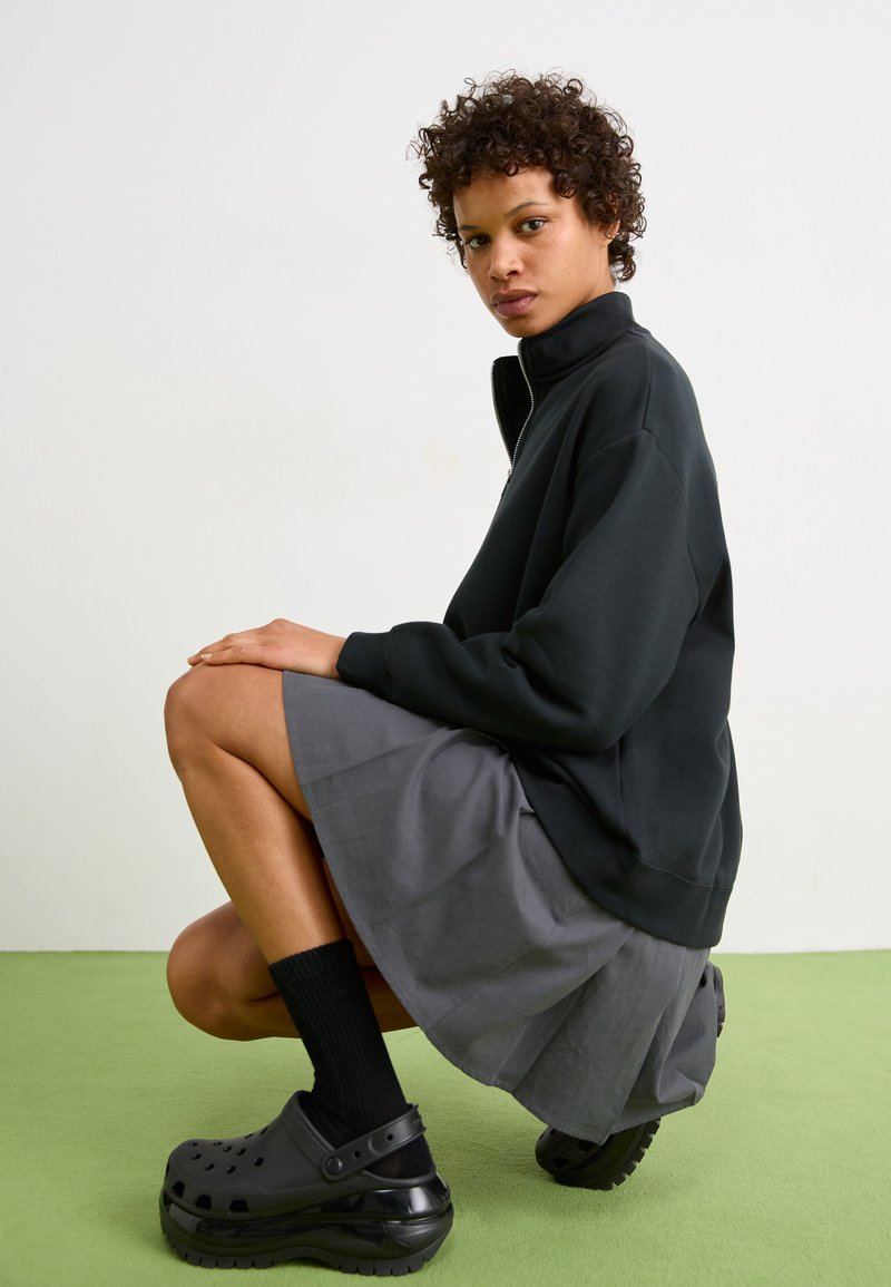 Sudadera negra con cremallera, falda gris con pliegues, zuecos de plataforma negros y calcetines oscuros. Modelo en cuclillas contra un fondo claro.