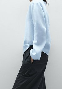 Henkilö, jolla on yllään vaalea sininen pitkähihaispaida ja kädet taskuissa, pukeutuneena väljät mustat housut, taustalla yksinkertainen vaaleanharmaa tausta.