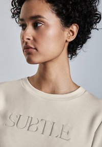 Krämfärgad sweatshirt med broderad textdesign "SUBTLE". Mjukt tyg med rund halsringning och slät textur.