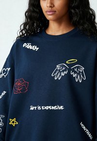 Marineblauwe sweatshirt met geborduurde motieven waaronder een rode roos, engelenvleugels en tekst: "Kunst is Duur" en "PANAM." Zachte textuur.