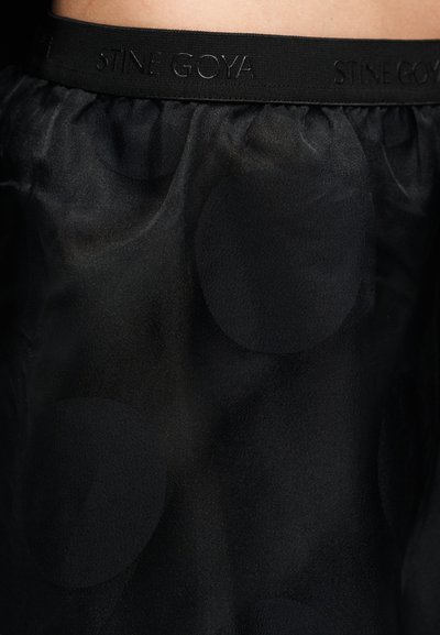 Svart genomskinlig kjol med mjuk textur och stort prickigt mönster, med en kontrasterande svart elastisk midjerem präglad med "STINE GOYA."