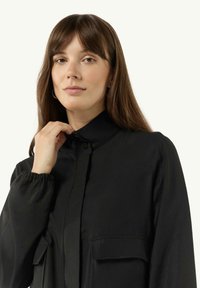 Czarna koszula z guzikami i wysokim kołnierzem, z elastycznymi mankietami oraz dwiema kieszeniami na piersi; gładka tekstura i minimalistyczny design.
