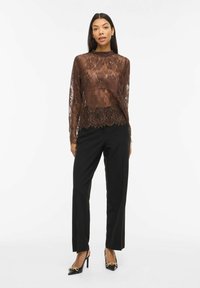 Femme portant un chemisier long à manches longues en dentelle marron transparente, un pantalon noir sur mesure et des talons pointus noirs avec détail chaîne dorée.