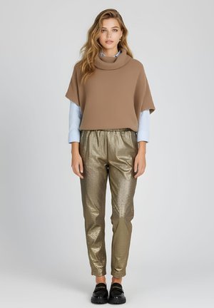 Joven mujer con un suéter marrón holgado de cuello alto, camisa azul clara, pantalones metálicos dorados y mocasines negros, de pie frente a un fondo liso.