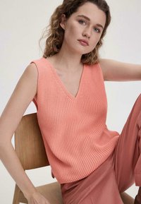 Helles korallenfarbenes, ärmelloses Stricktop mit V-Ausschnitt, strukturiertem Stoff und lockerer Passform, kombiniert mit passenden Hosen, sitzt auf einem Holzstuhl.