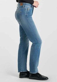 Jeans en denim bleu clair à coupe slim, avec une taille mi-haute, des poches arrière et une étiquette de marque en cuir marron à la taille.