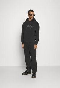 Felpa nera con tasca frontale, caratterizzata da testo bianco. Abbinate a pantaloni scuri e scarpe nere, insieme a occhiali da sole. Design semplice e moderno.