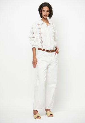 Witte blouse met kanten details, gecombineerd met witte high-waist jeans en een bruine riem. Geaccentueerd met tan gevlochten sandalen.