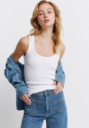 Junge Frau trägt ein weißes Tanktop und blaue Jeans, teilweise mit einer Jeansjacke drapiert, posiert vor einem einfarbigen weißen Hintergrund.