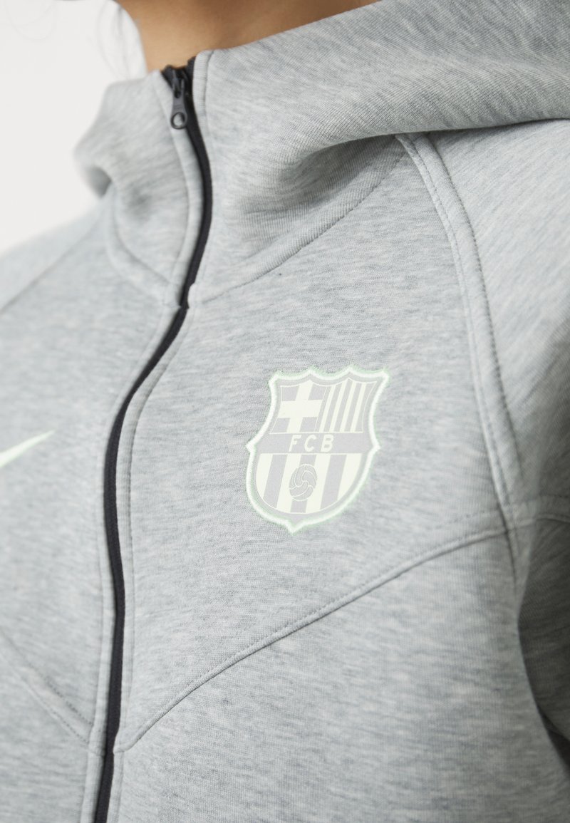 Zbliżenie szarej kurtki zapinanej na zamek z czarnym suwakiem i wyraźnym haftowanym logo klubu sportowego na piersi.