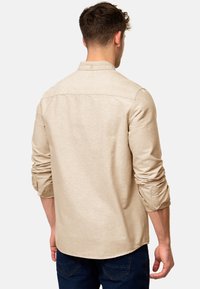 Chemise beige à manches longues avec col boutonné, manches retroussables et tissu texturé, associée à un jean bleu foncé.