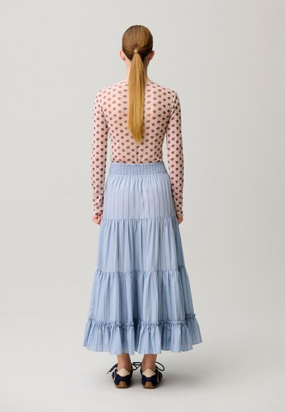 Donna con lunga coda di cavallo che indossa una maglia floreale a maniche lunghe, gonna a balze azzurro chiaro e scarpe nere, in piedi di spalle su sfondo semplice.