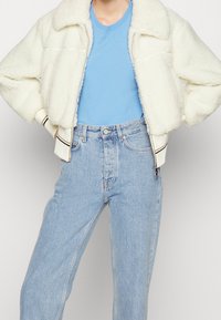 Person i ljusblå jeans, blå t-shirt och en kort vit fleecejacka med händerna på höfterna mot en enkel bakgrund.