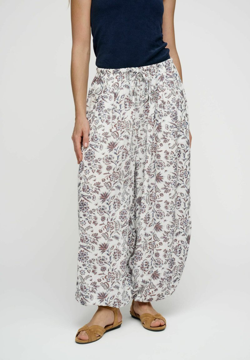 Femme portant un pantalon blanc ample imprimé floral avec taille à cordon, un haut sans manches bleu marine et des sandales beige, debout sur un fond uni.