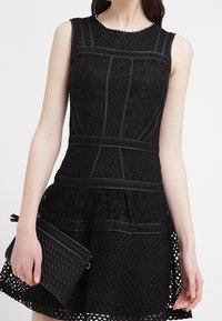 Svart spetsklänning med texturerade mönster, åtsittande bodice, utställd kjol och kontrasterande detaljer. Kombinerad med en svart flätad kuvertväska.