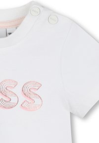 T-shirt en coton blanc à manches courtes arborant un logo "SS" brodé en rose et gris métallique, avec deux boutons sur l'épaule.