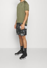 Zelené sportovní tričko s krátkým rukávem a logem, v kombinaci s černými kraťasy s maskáčem, které mají bílé logo. Černé sportovní boty a šedá láhev na vodu.